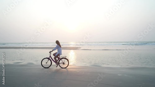 A young longhaired girl in a white dress riding a bike on the sandy tropical beach at sunset. Shooting with a drone.