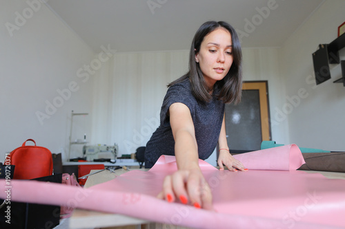 seamstress got high-quality material for bags from friend living abroad. Smiling girl with kare haircut and red nails unwrapping roll of leather on table in workshop at home. Concept of expen photo