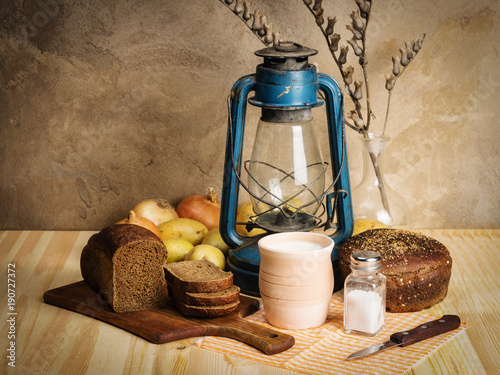 Pot of milk, rye bread, a kerosene lantern and vegetables on the table. Still-life in the rustic style