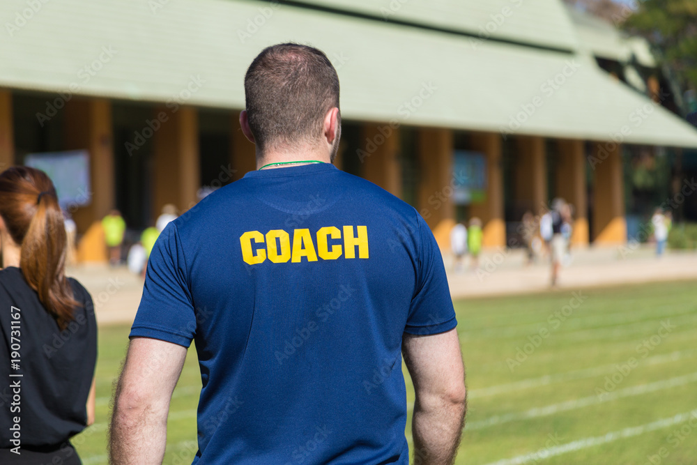 Back side of a male athlete coach in deep blue t-shirt with yellow COACH word written on its back Back side of a male athlete coach in deep blue t-shirt with yellow COACH word written on its back