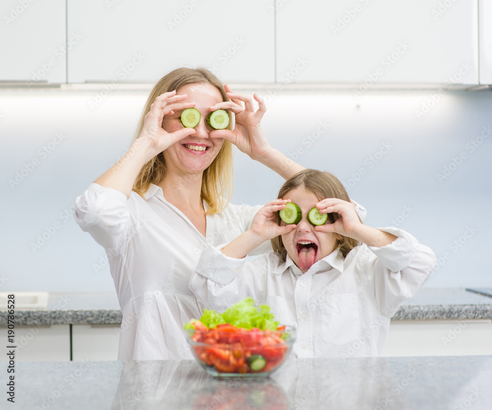 Happy mother and little girl having fun with food vegetables at kitchen holds cucumbers before his eyes like in glasses Happy mother and little girl having fun with food vegetables at kitchen holds cucumbers before his eyes like in glasses