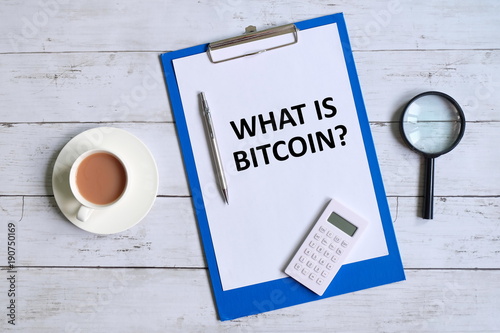 Top view of clipboard with paper written 'WHAT IS BITCOIN?' with pen,magnifying glass,calculator, and a cup of coffee on white wooden background.