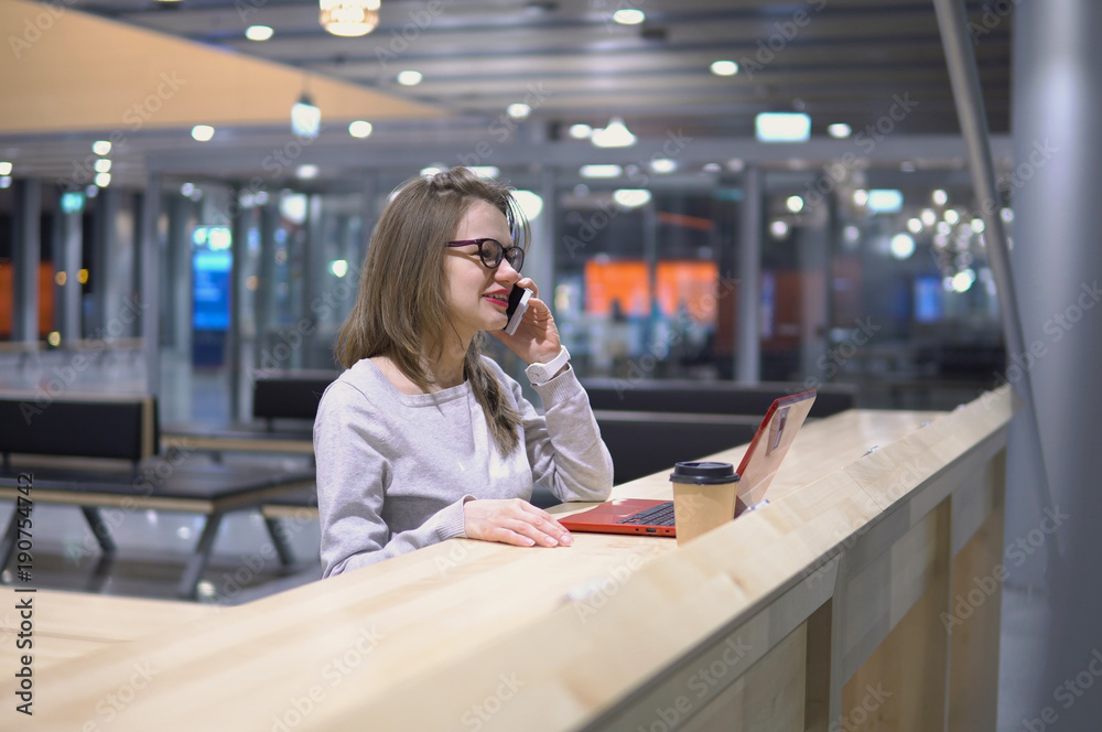 Young, beautiful girl talking on the phone standing at a table with a laptop and a cup of coffee at the airport