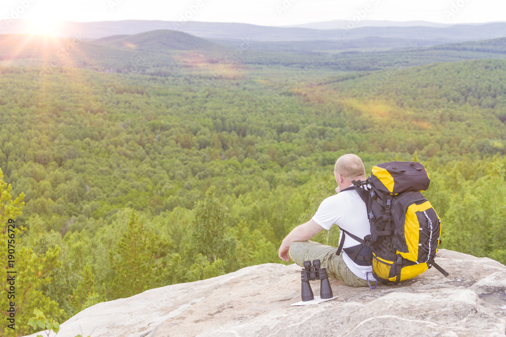 A tourist man is sitting with a backpack on top of a mountain, looking at the sunset. Camping, traveling, adventure.