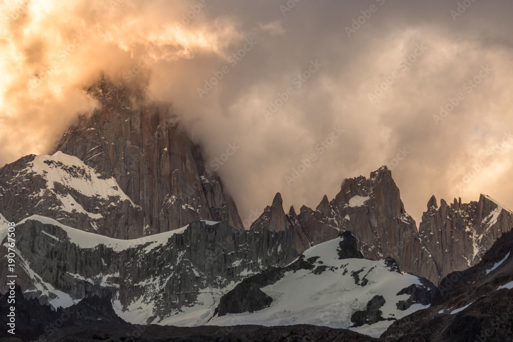 Patagonian adventure. On the background of a twilight sky, The Andes Mountains reveals their granite rock while are being surrounded of clouds. The highest peak is the Monte Fitz Roy.
