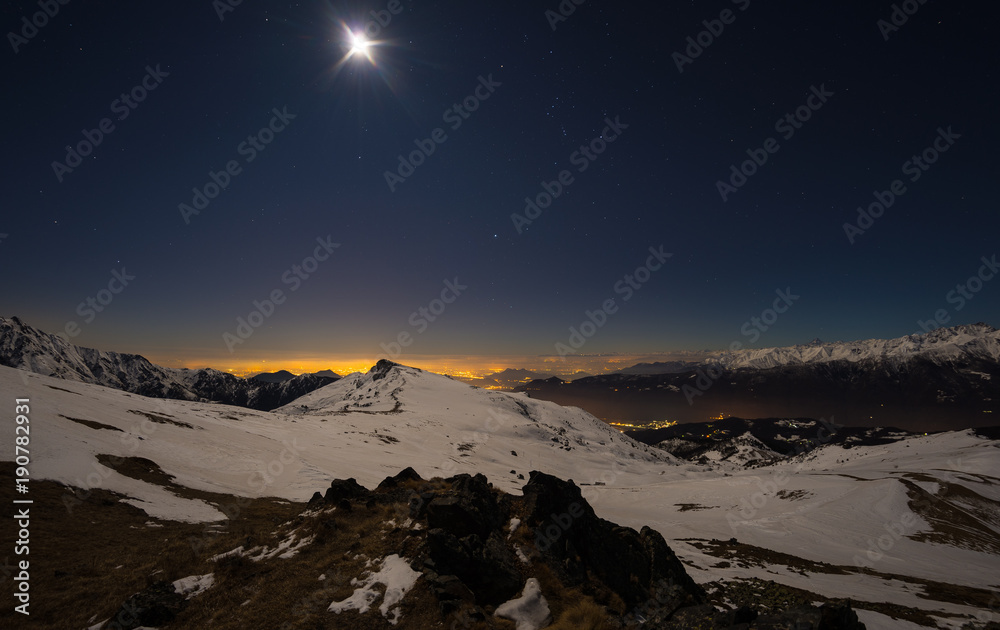 Turin city lights, night view from snow covered Alps by moonlight. Moon and Orion constellation, clear sky, fisheye lens. Italy.