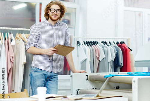 Young specialist of typography design with notepad standing by printing machine in studio