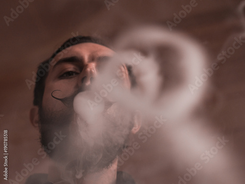 Man with Beard and Mustages Vaping an Electronic Cigarette. Vaper Hipster Smoke Vaporizer and Exhals Smoke Rings.