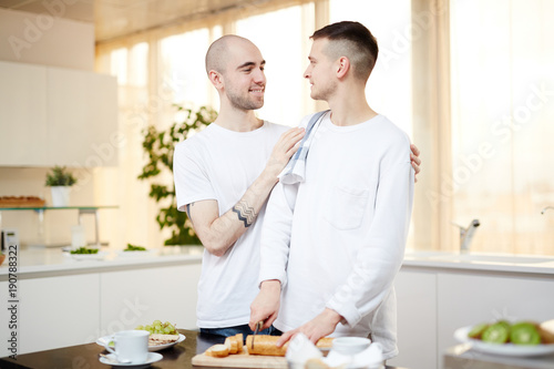 One of young homosexual men embracing his partner cutting bread for breakfast