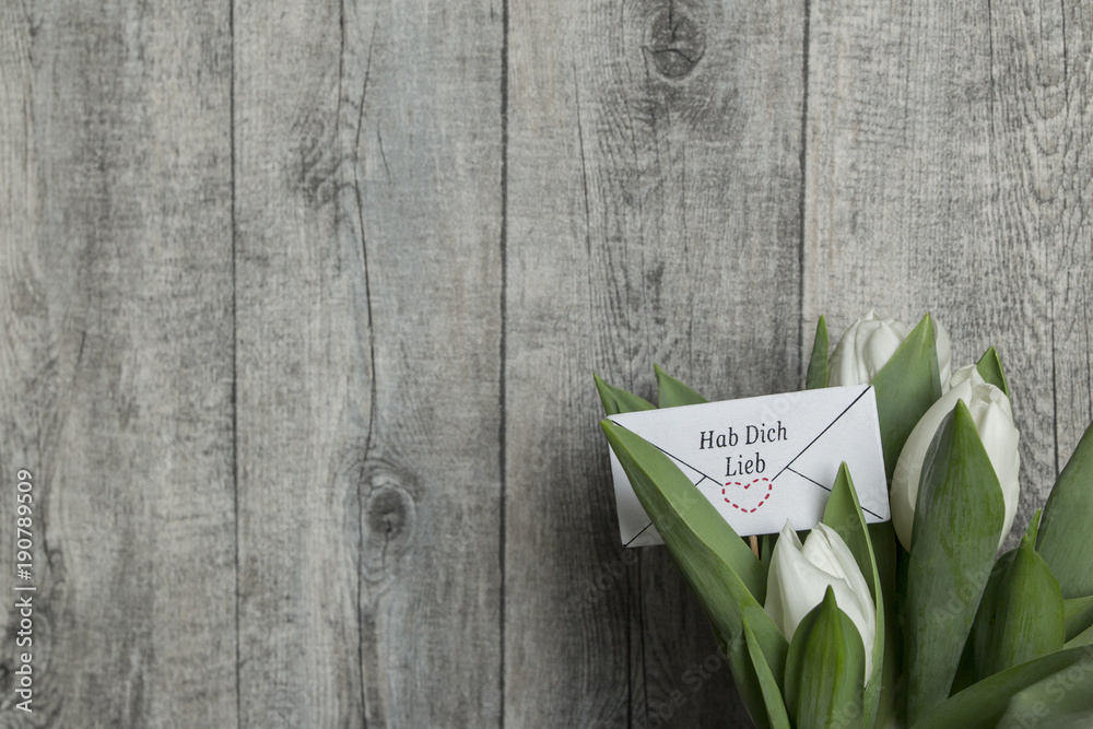 HAB DICH LIEB Brief mit Schriftzug und Herz in 3 weissen Tulpen vor grauer Holzwand Stock Photo