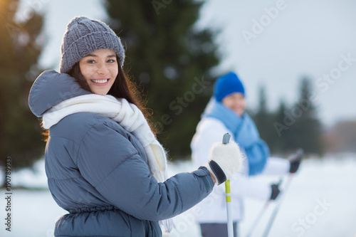 Smiling girl in grey beanie, white scarf and warm winter jacket looking at camera while skiing with her boyfriend in park or forest