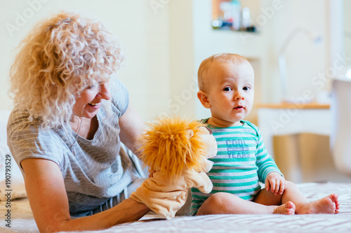 Blond curly female nanny playing with happy infant baby boy while sitting on the bed in cozy interior bedroom. Side view. Real people life and different generations concept. photo