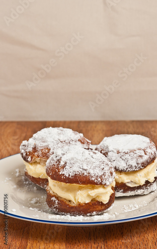 Three, freshly baked, homemade, white cream filled donuts with powder sugar, on a round, white plate with a blue rim on a wood serving board