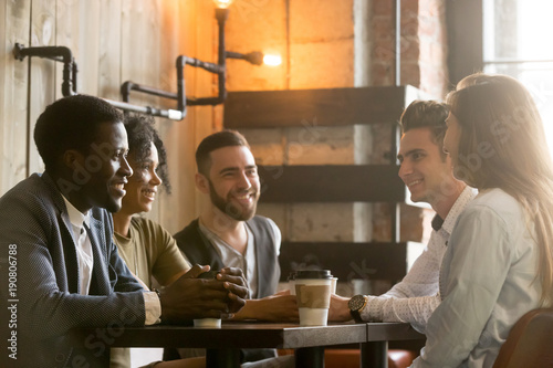 Multiracial happy friends talking and drinking coffee sharing coffeehouse table, diverse african and caucasian millennial young people smiling enjoying pleasant conversation in cafe at coffee break