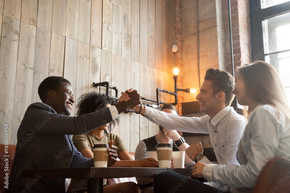 African american and caucasian men shaking hands at coffeehouse meeting with diverse friends, multiracial smiling male buddies greeting with friendship handshake sitting sharing table in cozy cafe