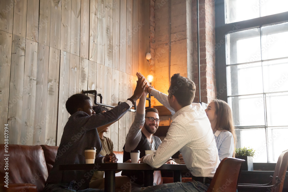 Fototapeta premium Multiracial best friends group giving high five sitting at cafe table, happy diverse african and caucasian young people join hands up together at meeting, unity and support in friendship concept