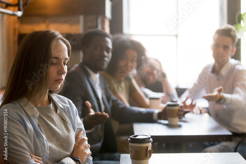 Multiracial friends trying to make peace with insulted girl offended after bad joke, diverse young people apologizing aggrieved resentful drama queen woman sitting apart sulking at meeting in cafe