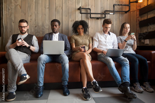 Diverse young people sitting in row on couch together obsessed with devices online, african and caucasian millennial addicts using laptops and smartphones, digital life and gadgets overuse concept