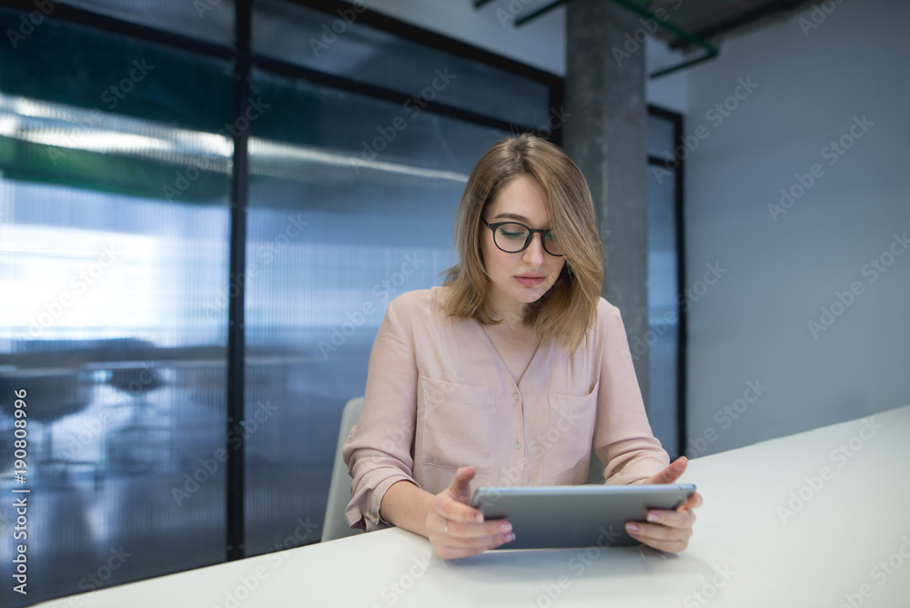 Beautiful young woman in glasses sitting at the desk in the office and using a tablet. Look at the tablet. Beautiful young woman in glasses sitting at the desk in the office and using a tablet. Look at the tablet.