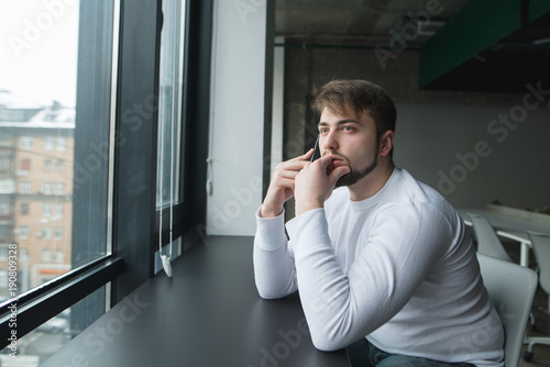 A young man with a beard sits in a modern office at the table near the window and talks on the phone. The man speaks on the phone and looks in the window