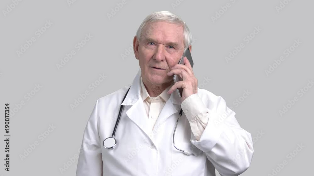 Portrait of smiling senior doctor talking on mobile phone. Old granddad working as doctor wearing stethoscope and talk on smartphone in grey isolated background.