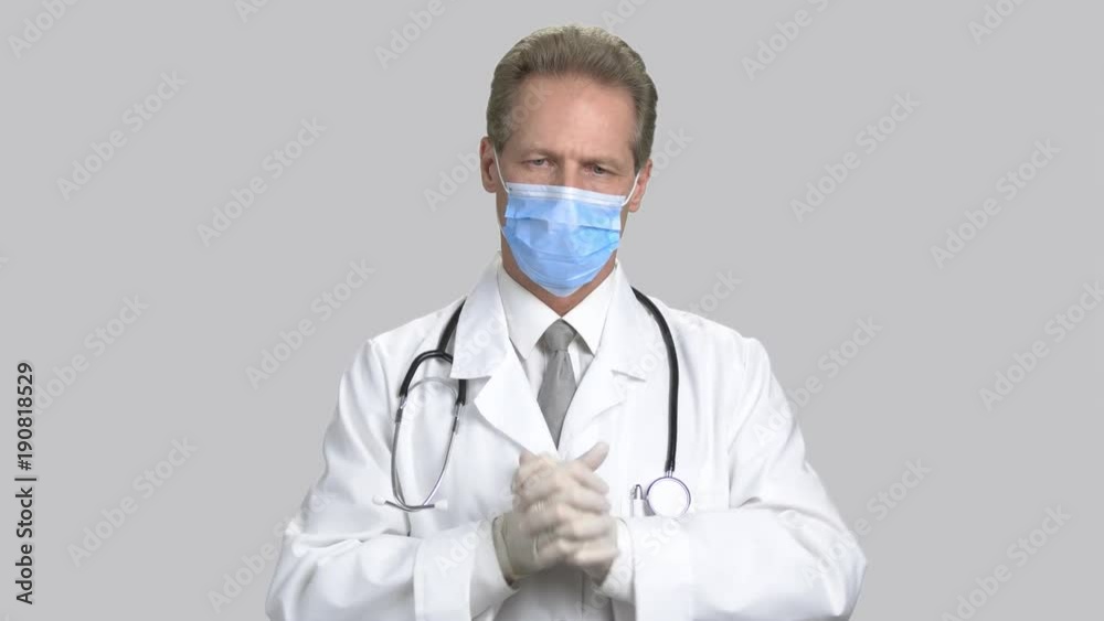 Portrait of doctor in mask put on gloves and smiling. Middle aged physician wear gloves, ready to operation. Grey isolated background. Portrait of doctor in mask put on gloves and smiling. Middle aged physician wear gloves, ready to operation. Grey isolated background.