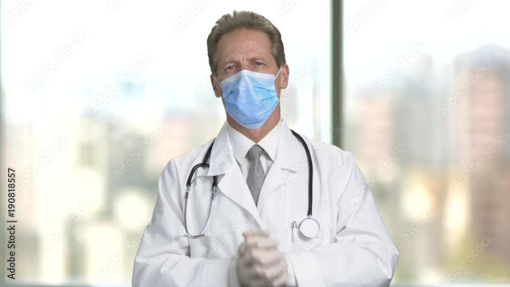 Portrait of doctor in mask fold hands and arms. Put on elastic rubber gloves. Bright abstract blurred windows background with view on city. Portrait of doctor in mask fold hands and arms. Put on elastic rubber gloves. Bright abstract blurred windows background with view on city.