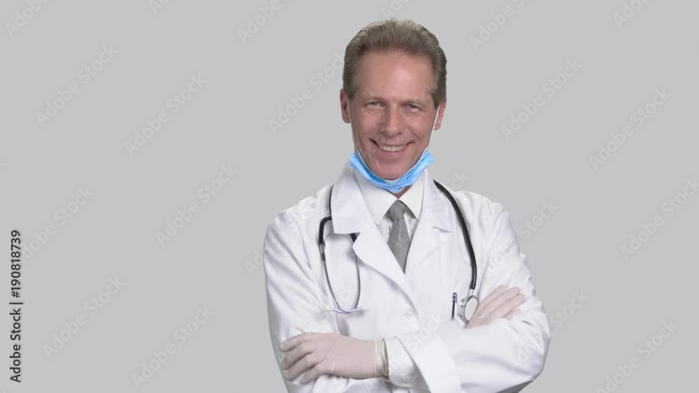 Mature doctor wearing medical gloves and folding arms. White medical suit with tie, stethoscope and mask wearing. Grey isolated background. Mature doctor wearing medical gloves and folding arms. White medical suit with tie, stethoscope and mask wearing. Grey isolated background.