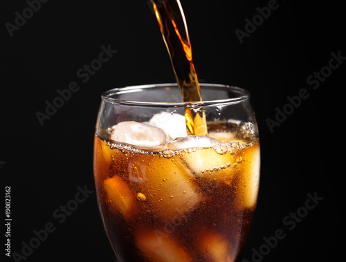 Pouring cola into glass with ice against black background, closeup