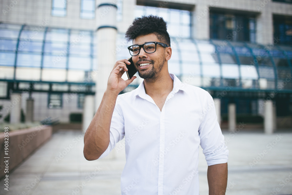 Positive and handsome Afro American man standing in front of huge modern business building smiling and talking on cell phone Positive and handsome Afro American man standing in front of huge modern business building smiling and talking on cell phone