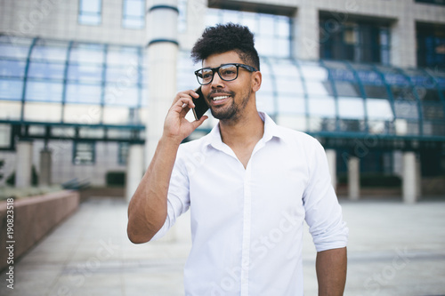 Positive and handsome Afro American man standing in front of huge modern business building smiling and talking on cell phone
