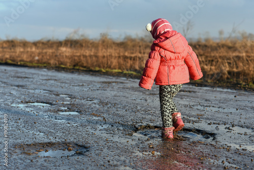 Little girl plays in puddle, children's fun, unforgettable moments, dirty and wet shoes, fun with father, life in village, sunlight. Concept - little roister. photo