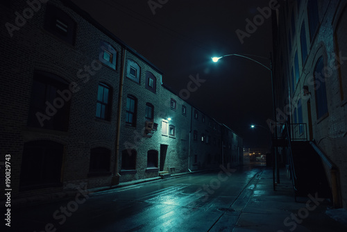 Dark urban city alley at night after a rain featuring vintage warehouses.