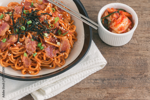 Korean hot and spicy instant noodle in blac bowl topped with bacon, sesame, seaweed and chopped scallion served with kimchi on wood table with copy space. Homemade delicious asian food concept.