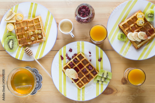 Homemade Belgian waffles with jam, honey, pieces of banana, kiwi fruit, kumquat and cranberries on striped plates and a glass of espresso and peach juices in glasses on a wooden table.