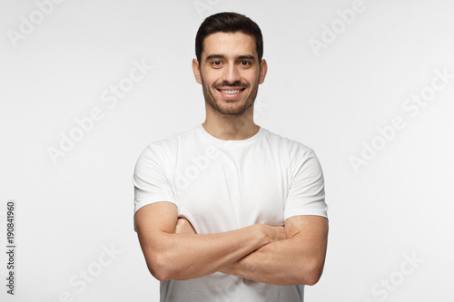 Portrait of smiling handsome man in white t-shirt standing with crossed arms isolated on gray background