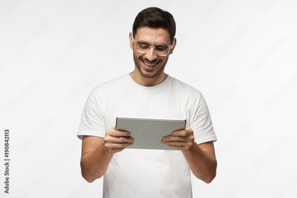 Studio closeup of young man pictured isolated on grey background wearing transparent eyeglasses looking attentively to screen of tablet he is holding, smiling happily, laughing Studio closeup of young man pictured isolated on grey background wearing transparent eyeglasses looking attentively to screen of tablet he is holding, smiling happily, laughing