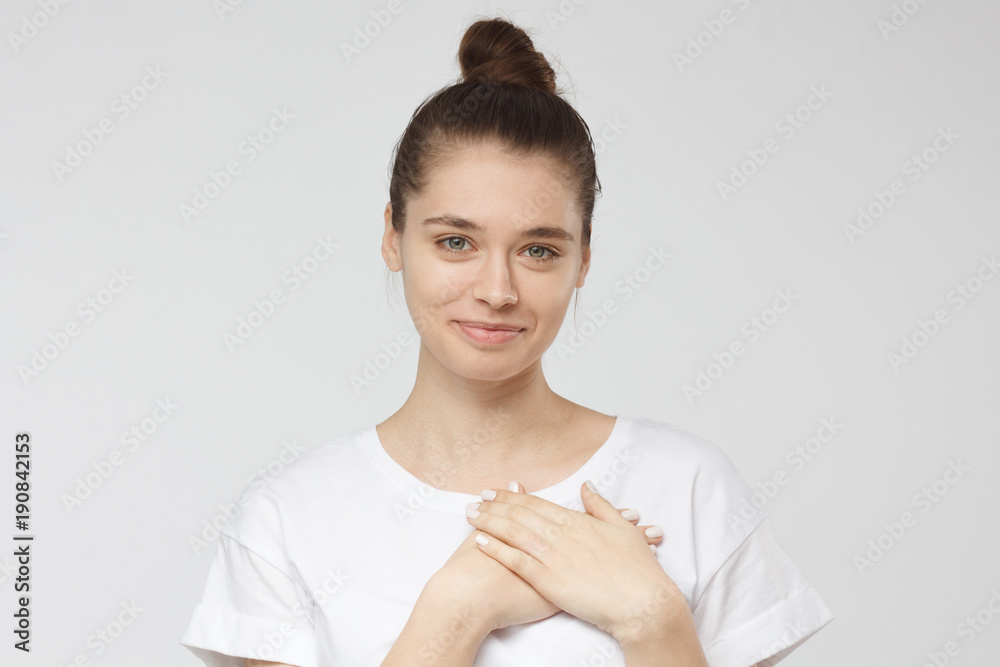 Portrait of smiling positive young woman keeps hands on chest, expresses sympathy. Kind hearted friendly nice girl shows kindness, wearing blank white t-shirt Portrait of smiling positive young woman keeps hands on chest, expresses sympathy. Kind hearted friendly nice girl shows kindness, wearing blank white t-shirt
