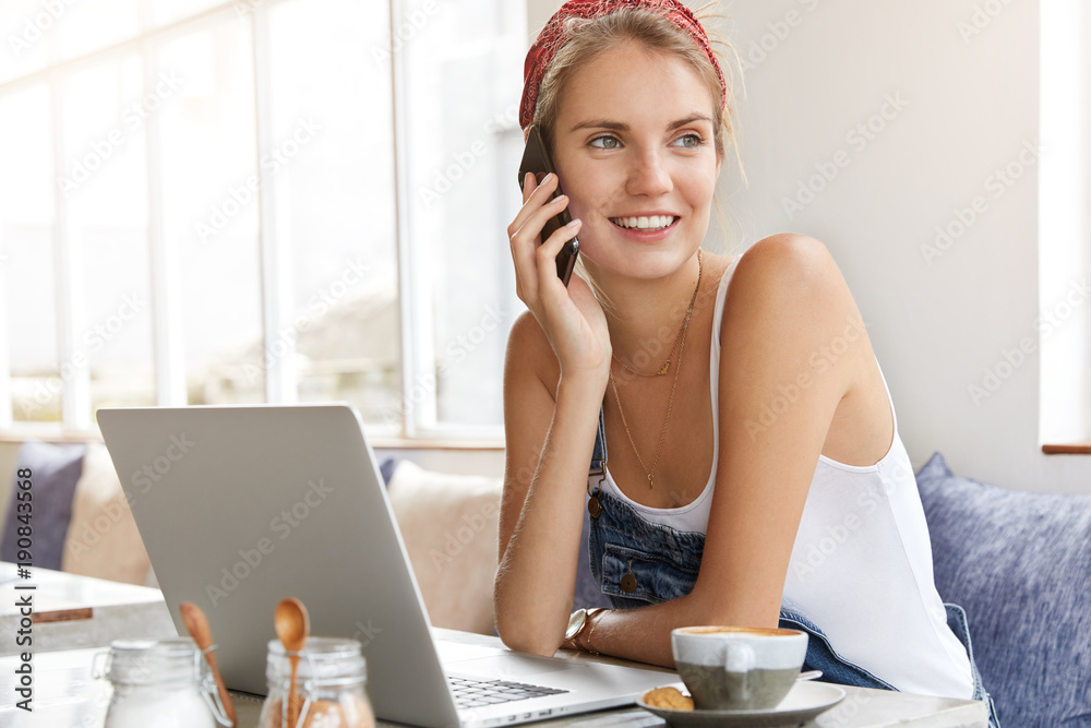 Obraz premium Skilled copywriter or freelancer dressed casually, has pleasant mobile conversation, expresses positiveness and happiness, works distantly on laptop computer, enjoys strong coffee or hot tea