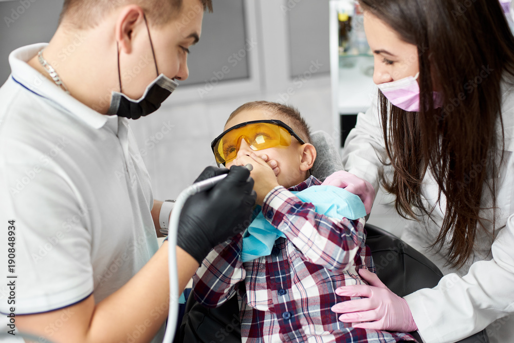 Fototapeta premium A child in orange dental glasses is covering his mouth being scared before his dental treatment being performed by a young dentist and his attractive female assistant.