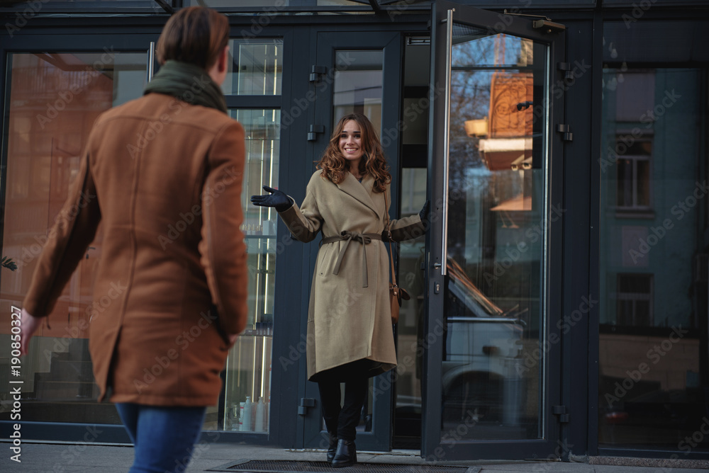 Fototapeta premium Joyful young woman is going out the door of building while meeting her boyfriend. She is looking at man with happiness and smiling