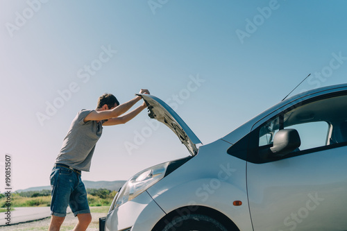 Young man looking under the hood of breakdown car outdoors. Transportation and vehicle concept