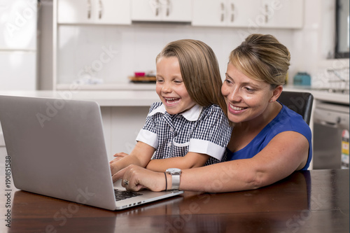 blond mother woman together with her young beautiful and sweet little girl 6 to 8 years old sitting at home kitchen enjoying with laptop computer