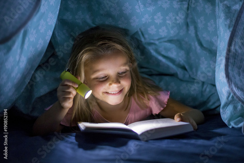 sweet beautiful and pretty little blond girl 6 to 8 years old under bed covers reading book in the dark at night with torch light smiling happy