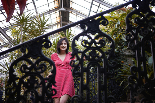 Portrait of fashionable European teenage girl with dark bob haircut having fun in plant nursery surrouned by various exotic trees and smiling happily at camera through patterned metal fence