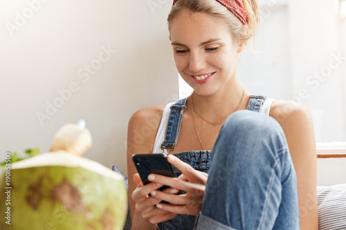 Positive female blogger rests at home, feels relaxed, texts message on mobile phone, uses modern application, writes information on website, enjoys exotic cocktail. People, technology and rest photo