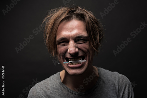 Portrait of grubby cheerful guy clutching syringe between teeth with happy grin. Isolated on background