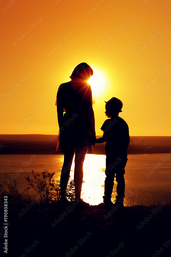 Silhouettes of mom and son holding hands and admiring the beautiful view of the sea on sunny evening at sunset time
