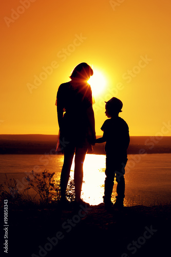 Silhouettes of mom and son holding hands and admiring the beautiful view of the sea on sunny evening at sunset time