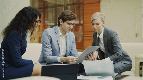 Businessman in glasses talking and duscussing future contract with female business partners sitting on couch in meeting room photo